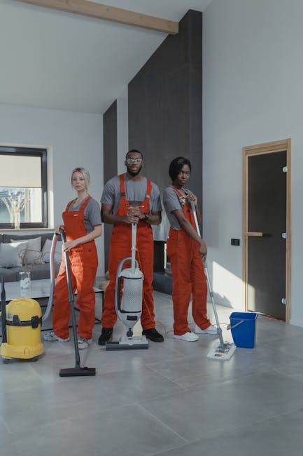 Three professional cleaners from Cleaner Shoreditch, wearing grey shirts and bright orange overalls, are standing on a modern, well-lit living room floor with large grey tiles. They are equipped with 
