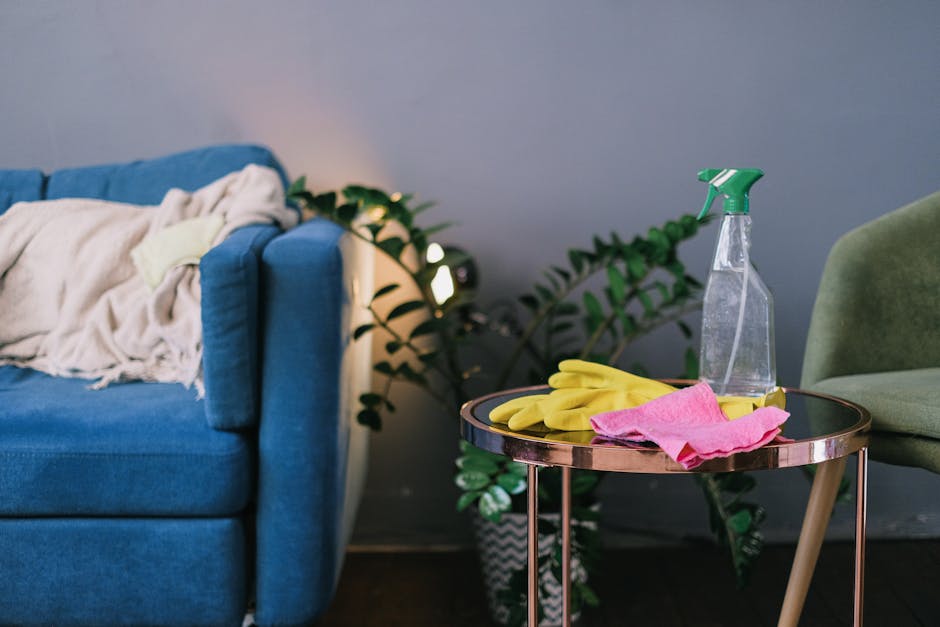Three professional cleaners from Cleaner Shoreditch, wearing grey shirts and bright orange overalls, are standing on a modern, well-lit living room floor with large grey tiles. They are equipped with 